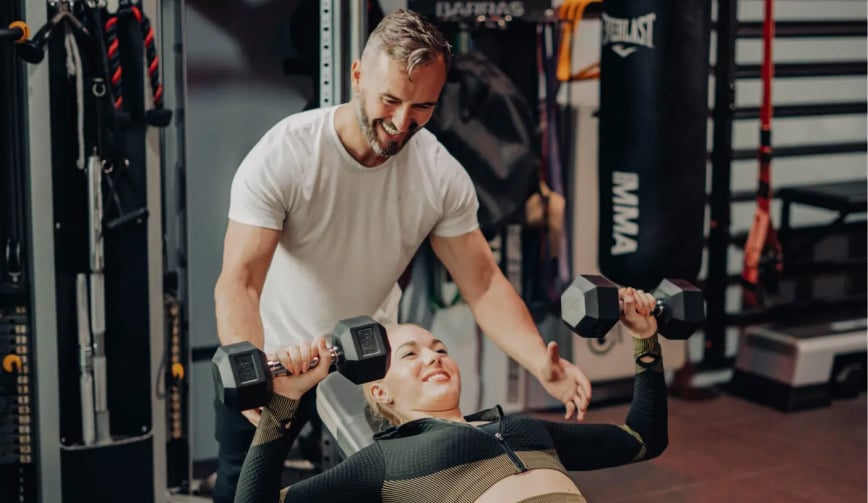 Entrenador personal supervisando a una mujer mientras realiza un press de banca con mancuernas en el gimnasio boutique Trim.